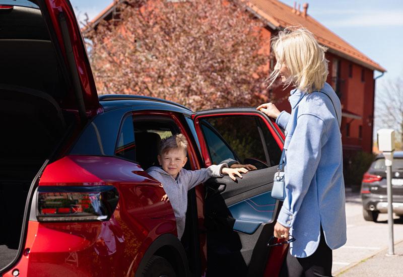 Lapsi katsoo pysäköitynä olevasta punaisesta Volkswagenista ulos astuessaan kameraan, kun nainen avaa hänelle oven.